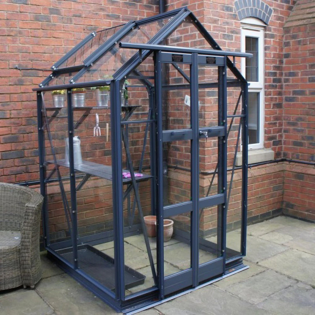 Angled view of Cottage 4 greenhouse in graphite finish, set against a brick wall with interior shelving and potted plants