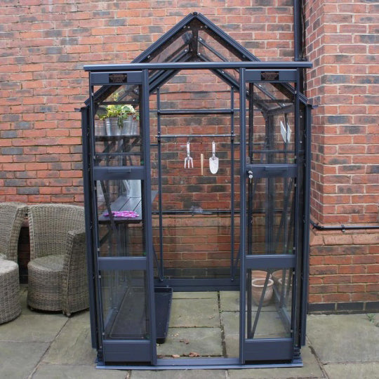 Front view of Cottage 4 greenhouse in graphite black finish, staged against a brick wall with tools hanging inside
