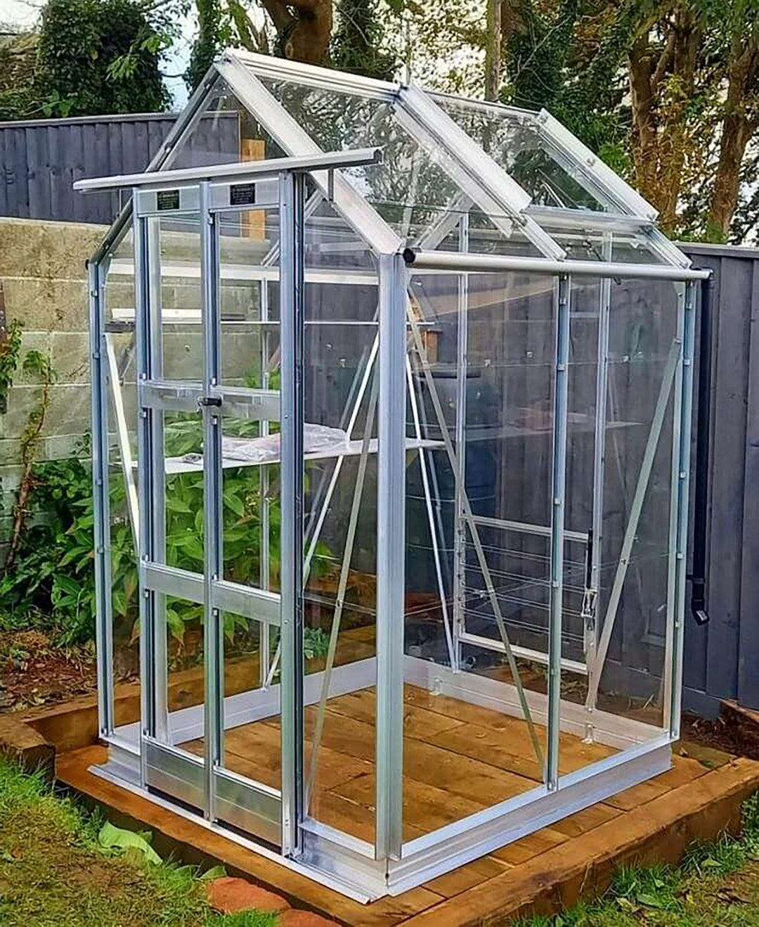 Cottage 4 greenhouse in natural aluminum finish with toughened glass, installed on a wooden platform in a shaded garden