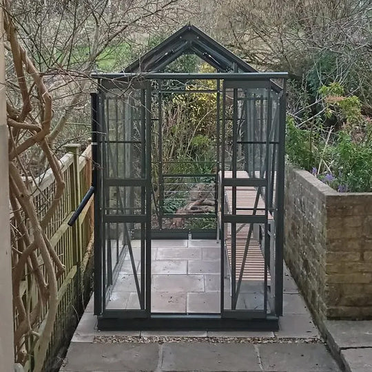 Front view of Cottage 8 greenhouse in graphite finish, situated between fence and wall on a tile base