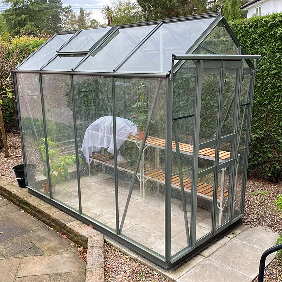 Cottage 8 greenhouse in green finish with two-tier staging, shown in a landscaped backyard on a stone base